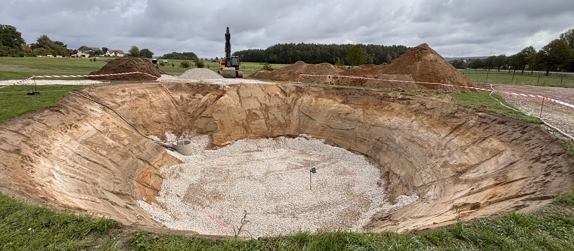 Kreisrunde Baugrube mit Kiesbett auf einer Wiese, dahinter ein Bagger und Aushubhügel